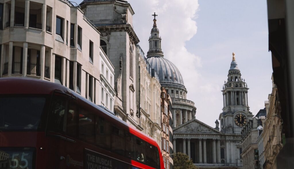 View of St Paul's Cathedral dome and City of London street scene, with a red London double-decker bus in the foreground and pedestrians walking near the cathedral steps. Cover image for blog on offshore bonds and Lombard lending.